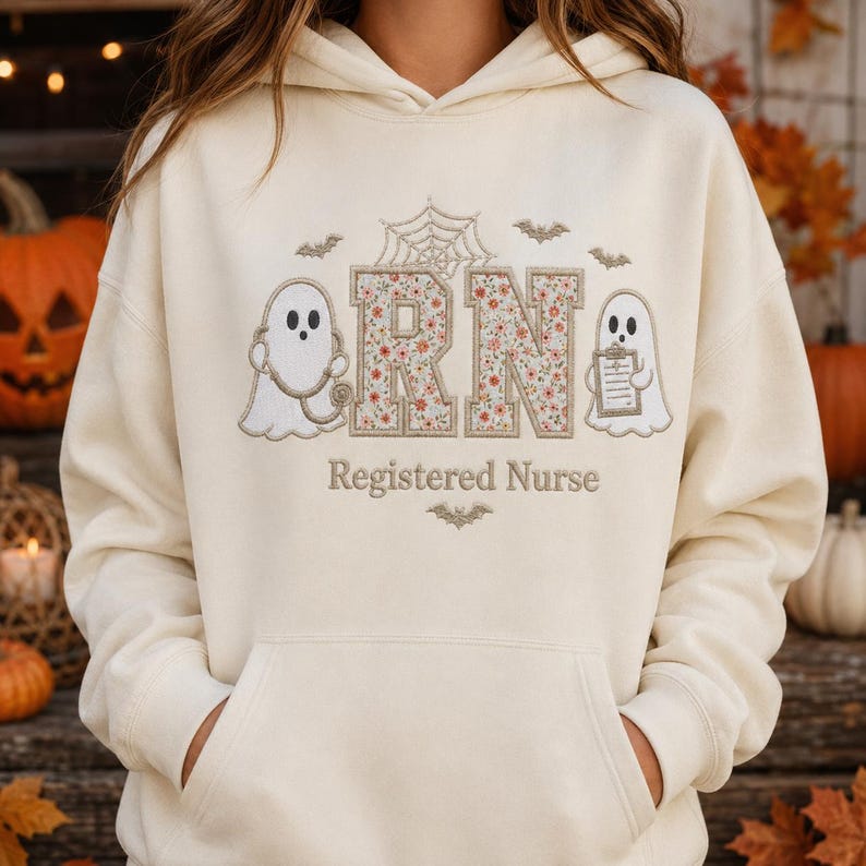 May include: A cream-colored hooded sweatshirt featuring an embroidered design with two white ghosts, a spider web, bats, and the letters RN filled with a floral pattern. Below the letters, the text Registered Nurse is embroidered in brown.