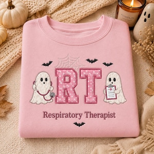May include: A light pink crewneck sweatshirt featuring embroidered designs of two white ghosts, one holding a stethoscope and the other a clipboard. The letters RT are in pink with white polka dots, accompanied by a spiderweb, bats, and the text Respiratory Therapist.