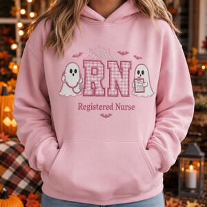 May include: A light pink hooded sweatshirt featuring an embroidered design with the letters RN in a pink and white gingham pattern. The design includes two white ghosts, one holding a stethoscope and one holding a clipboard, with spiderwebs and bats.