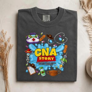 May include: A charcoal gray t-shirt featuring a graphic print with the text CNA STORY TO INFINITY AND BEYOND FOR OUR PATIENTS. The design includes a medical kit, stethoscope, cowboy hat, bandage, rocket, medicine bottle, and a face mask.