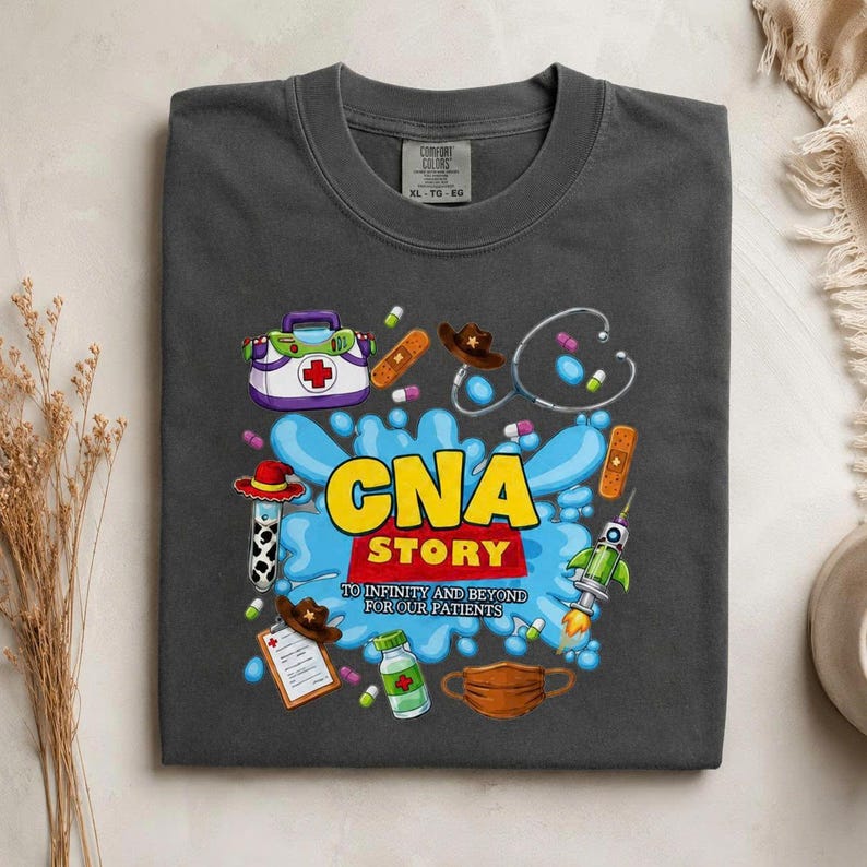 May include: A charcoal gray t-shirt featuring a graphic print with the text CNA STORY TO INFINITY AND BEYOND FOR OUR PATIENTS. The design includes a medical kit, stethoscope, cowboy hat, bandage, rocket, medicine bottle, and a face mask.