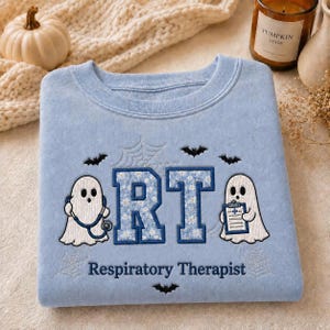 May include: A light blue crewneck sweatshirt featuring embroidered white ghosts, black bats, and spiderwebs. The center displays the letters RT in a blue floral pattern, with the text Respiratory Therapist below. One ghost holds a stethoscope.