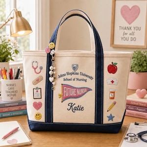 May include: A cream-colored canvas tote bag with navy blue trim, featuring embroidered medical icons, a pennant reading Future Nurse, and the text Johns Hopkins University School of Nursing with the name Katie. A beaded charm hangs from the handle.