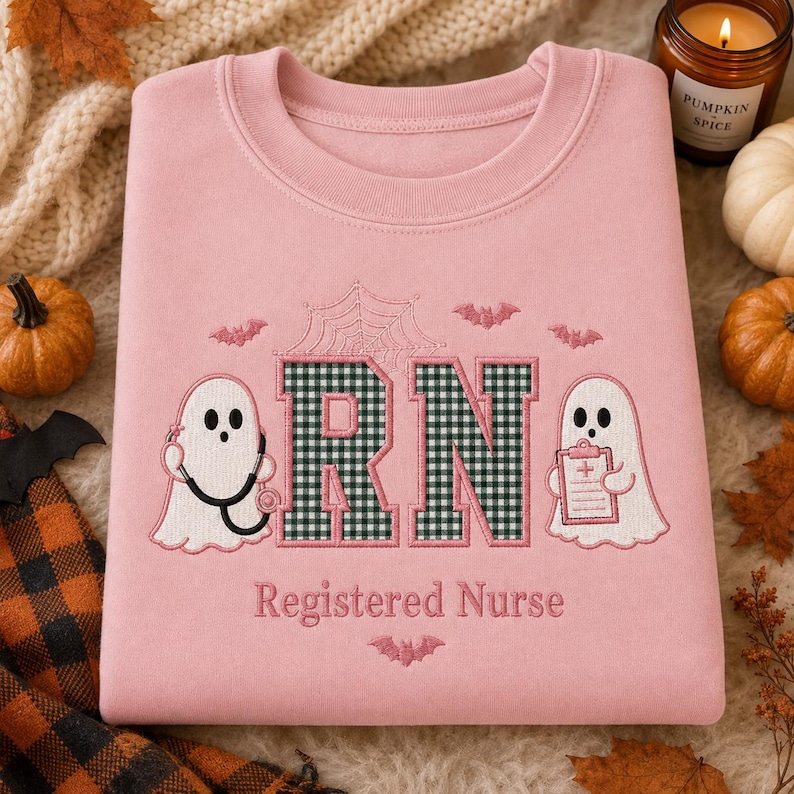 May include: A light pink crewneck sweatshirt featuring embroidered designs. The center displays the letters RN in a dark green and white gingham pattern, flanked by two white ghosts, one holding a stethoscope and the other a clipboard.