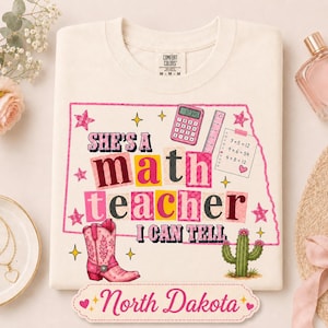May include: A cream-colored t-shirt featuring a pink outline of North Dakota. The graphic includes the text 'SHE'S A math teacher I CAN TELL', a pink calculator, a ruler, a math worksheet, a pink cowboy boot, and a green cactus.