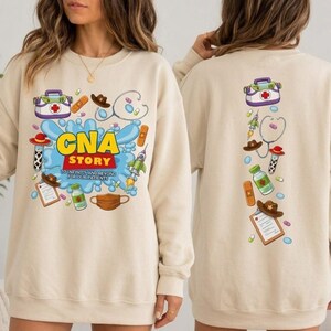 May include: A beige crewneck sweatshirt featuring a graphic print with the text 'CNA STORY TO INFINITY AND BEYOND FOR OUR PATIENTS' surrounded by medical and space-themed illustrations including a first aid kit, stethoscope, and rocket.