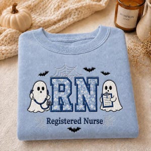 May include: A light blue crewneck sweatshirt featuring embroidered white ghosts, black bats, and spiderwebs. The center displays the letters RN in a blue floral pattern, with the text Registered Nurse below, accompanied by a stethoscope and clipboard.