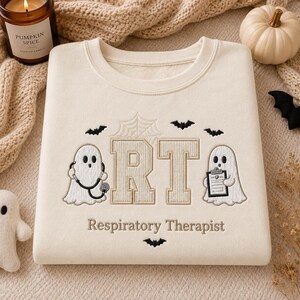 May include: A cream-colored crewneck sweatshirt featuring embroidered designs including two white ghosts, one holding a stethoscope and the other a clipboard, the letters RT, a spiderweb, bats, and the text Respiratory Therapist.