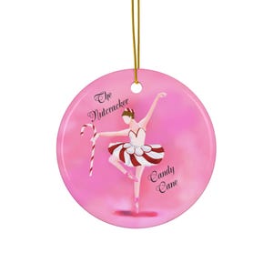 Op de afbeelding: Een rond roze keramieken ornament met een ballerina in een wit en rood gestreepte tutu die een zuurstok vasthoudt. De tekst "The Nutcracker" staat boven de danser en "Candy Cane" staat eronder, opgehangen aan een gouden koordje.