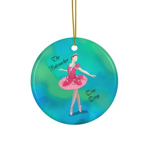 May include: Round ceramic ornament featuring a ballerina in a pink tutu and a matching bodice. The ornament has a gold string for hanging and the words "The Nutcracker" and "Dew Drop" are written on the ornament.
