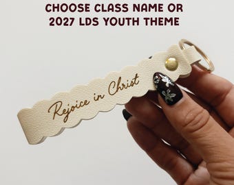 May include: A cream-colored scalloped keychain with a gold metal snap and ring. The text "Rejoice in Christ" is engraved on the front. Above the keychain, the text reads "CHOOSE CLASS NAME OR 2027 LDS YOUTH THEME".