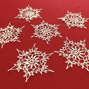 May include: Six white, crocheted snowflakes are arranged on a red background. Each snowflake has a detailed, openwork design with a central circular pattern and pointed edges. The snowflakes are a delicate, handmade craft.