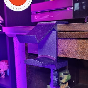 May include: A black 3D printed clamp with a screw, mounted to a wooden surface. The clamp is designed to hold a gaming console or other device. The clamp is in focus, while the background is blurred.