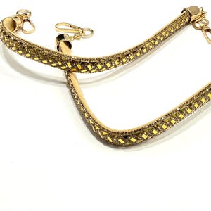 May include: A gold-colored purse strap with a geometric pattern of small, sparkling embellishments. The strap has gold-tone metal clasps on each end for attachment. The background is plain white.
