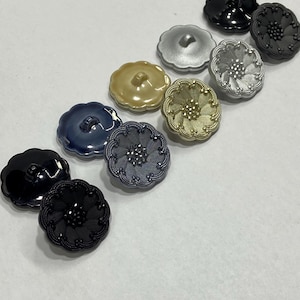 Op de afbeelding: Assortiment van acht decoratieve knopen met een bloemmotief. De knopen zijn in verschillende kleuren, waaronder zwart, zilver, goud en blauw.