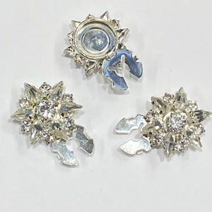 May include: Three silver-toned metal embellishments with clear rhinestone accents. Each embellishment has a starburst design and a clasp on the back.