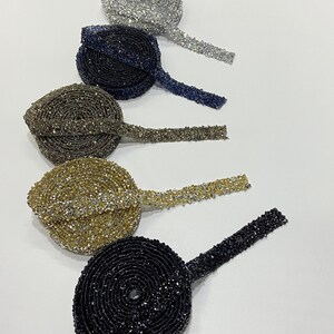 May include: Five spools of beaded trim in different colors. The trim is made of small, sparkly beads and is available in silver, navy blue, brown, gold, and black.