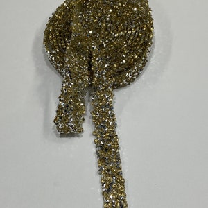 May include: A gold and silver rhinestone trim with a sparkly finish. The trim is rolled up and a long piece is hanging down.