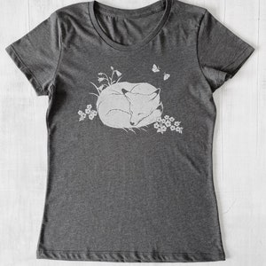 K&ouml;nnte beinhalten: Ein dunkelgraues T-Shirt mit einem wei&szlig;en Aufdruck eines schlafenden Fuchses, der sich in einem Blumenfeld zusammengerollt hat. Zwei Schmetterlinge fliegen &uuml;ber dem Fuchs.