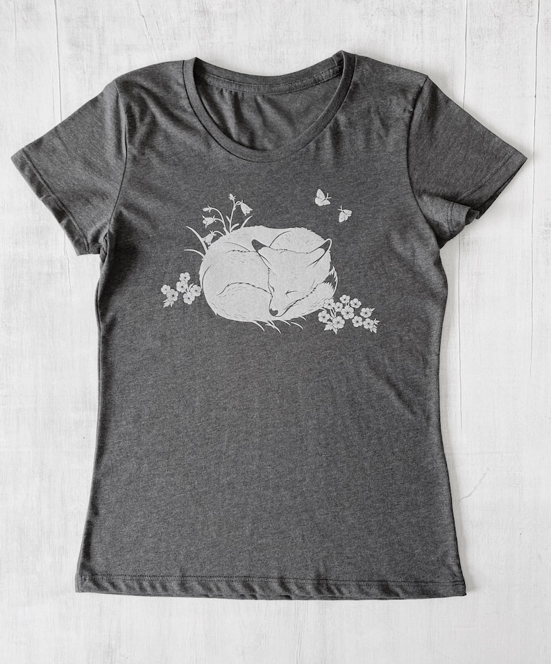 K&ouml;nnte beinhalten: Ein dunkelgraues T-Shirt mit einem wei&szlig;en Aufdruck eines schlafenden Fuchses, der sich in einem Blumenfeld zusammengerollt hat. Zwei Schmetterlinge fliegen &uuml;ber dem Fuchs.