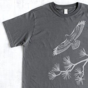 Może przedstawiać: Szary t-shirt z białym siluetem jastrzębia lecącego nad gałęzią sosny.