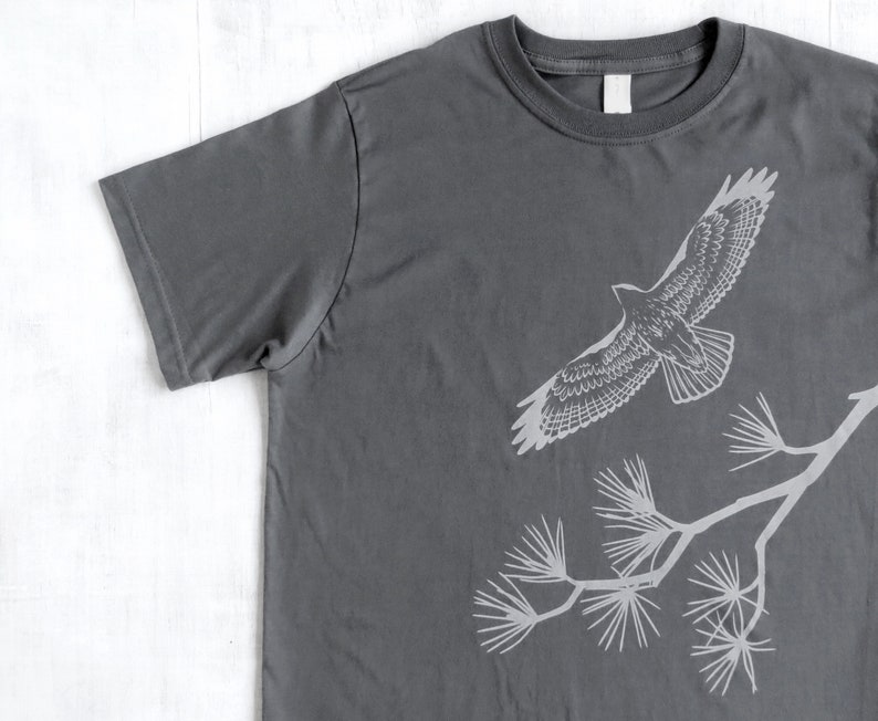 Może przedstawiać: Szary t-shirt z białym siluetem jastrzębia lecącego nad gałęzią sosny.