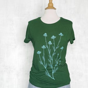 May include: A dark green short-sleeved t-shirt featuring a light blue graphic print of a cluster of chamomile flowers with thin stems and delicate leaves centered on the front of the garment, displayed on a mannequin torso.