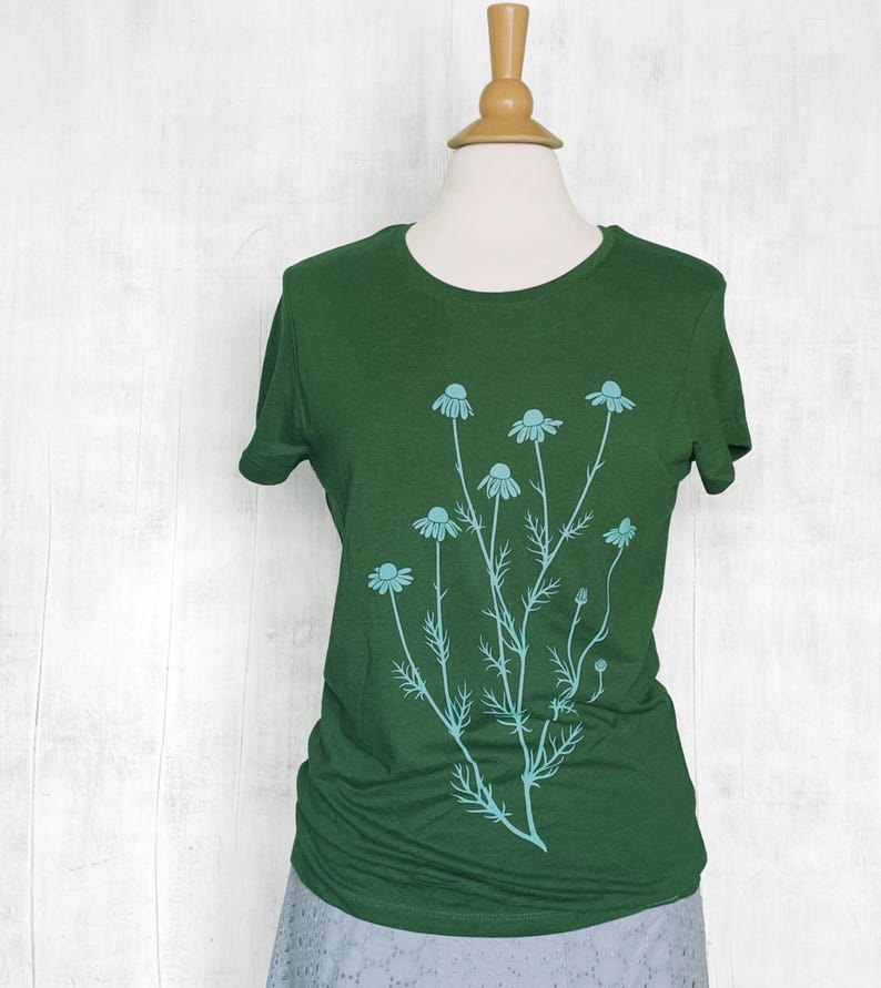May include: A dark green short-sleeved t-shirt featuring a light blue graphic print of a cluster of chamomile flowers with thin stems and delicate leaves centered on the front of the garment, displayed on a mannequin torso.