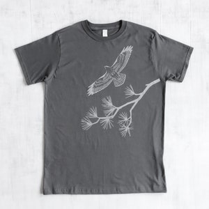 Może przedstawiać: Szary t-shirt z białym siluetem jastrzębia lecącego nad gałęzią z igłami sosny.