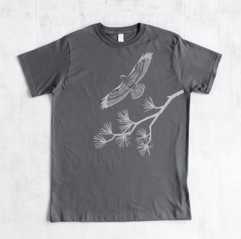 Może przedstawiać: Szary t-shirt z białym siluetem jastrzębia lecącego nad gałęzią z igłami sosny.