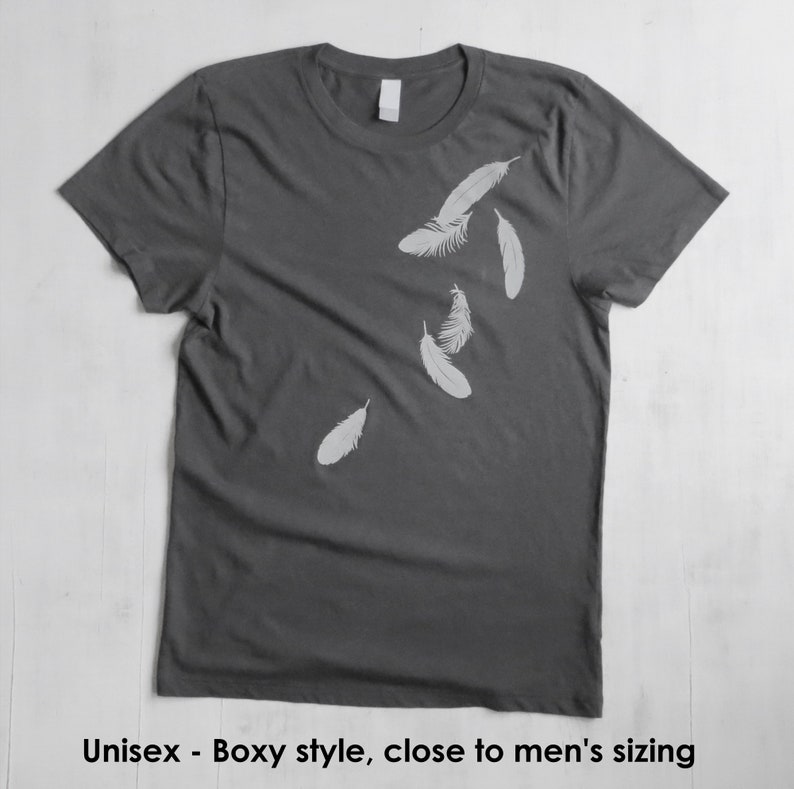 May include: A dark gray t-shirt with a white graphic of feathers falling. The shirt has a boxy fit.