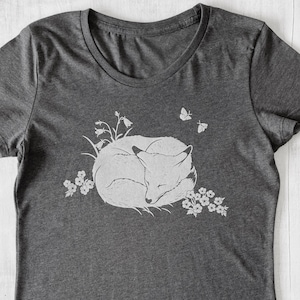 K&ouml;nnte beinhalten: Ein dunkelgraues T-Shirt mit einem wei&szlig;en Aufdruck eines schlafenden Fuchses, umgeben von Blumen und Schmetterlingen.