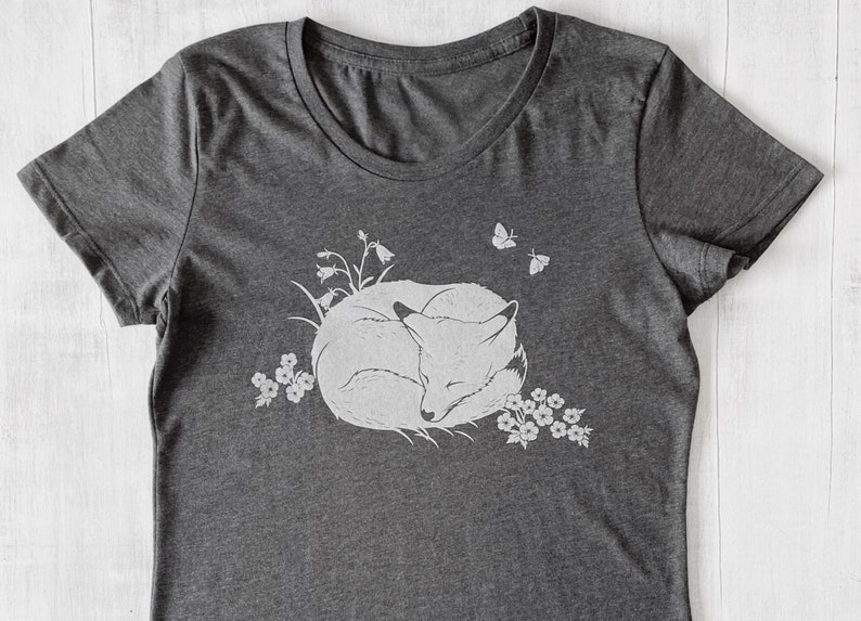 K&ouml;nnte beinhalten: Ein dunkelgraues T-Shirt mit einem wei&szlig;en Aufdruck eines schlafenden Fuchses, umgeben von Blumen und Schmetterlingen.