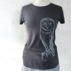 May include: A dark gray t-shirt with a white outline of an owl on the front. The owl is facing forward with its wings spread out.