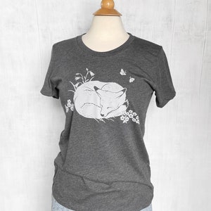 K&ouml;nnte beinhalten: Ein dunkelgraues T-Shirt mit einem wei&szlig;en Aufdruck eines zusammengerollten schlafenden Fuchses, umgeben von Blumen und Schmetterlingen.
