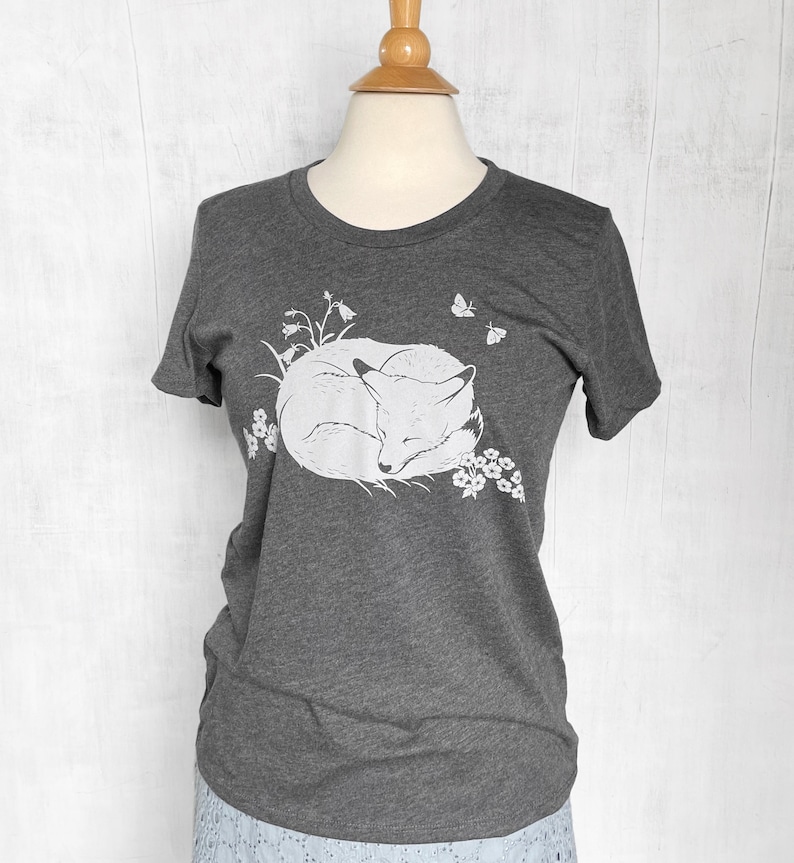 K&ouml;nnte beinhalten: Ein dunkelgraues T-Shirt mit einem wei&szlig;en Aufdruck eines zusammengerollten schlafenden Fuchses, umgeben von Blumen und Schmetterlingen.