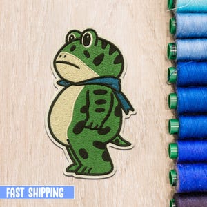 Puede incluir: Parche bordado con una rana verde con manchas negras y una bufanda azul. La rana tiene una expresión neutra y está de pie. El parche está sobre una superficie de madera clara con carretes de hilo azul.