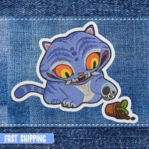 May include: Embroidered patch featuring a blue cat-like creature with orange eyes and sharp teeth. The patch includes a small overturned pot with a plant. The patch is on a denim background. The text "FAST SHIPPING" is at the bottom.