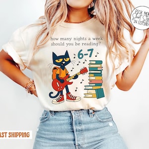 May include: Cream-colored t-shirt featuring a cartoon cat playing a red guitar in front of a stack of books. Text on the shirt reads "how many nights a week should you be reading?" and "6-7".