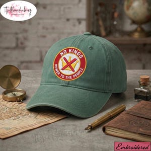 May include: A sage green baseball cap with a circular embroidered patch. The patch features the text "NO KINGS" and "POWER TO THE PEOPLE" around a red circle with a gold crown and a red "X" in the center. The cap is on a table.