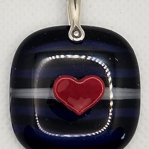 May include: A square glass pendant with a silver bail. The pendant has a dark blue background with white and black stripes. A red heart is in the center of the pendant.