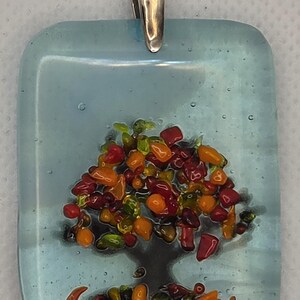 Puede incluir: Un colgante rectangular de vidrio fusionado con un fondo azul claro y la silueta de un árbol oscuro. La copa del árbol y el suelo están decorados con pequeñas piezas de vidrio multicolor en tonos rojos, naranjas, amarillos y verdes. Una anilla de metal plateado está fijada en la parte superior.