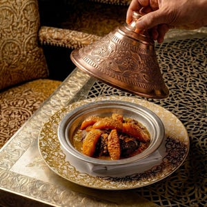 Op de afbeelding: Een koperen tajine-deksel wordt opgetild om een gerecht met eten te onthullen. Het eten wordt geserveerd in een zilveren kom op een goudgerande bord. Het gerecht bevat gekookte groenten en vlees. De achtergrond heeft sierlijke gouden en bruine patronen.