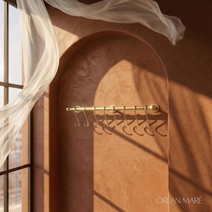 Może przedstawiać: Złoty metalowy wieszak na przybory kuchenne z siedmioma haczykami, zamontowany na teksturowanej, czerwonobrązowej ścianie. Wieszak umieszczony jest pod łukowatą wnęką. Przezroczysta biała tkanina układa się w pobliżu okna, a widoczna jest nazwa marki "ORLAN MARE".