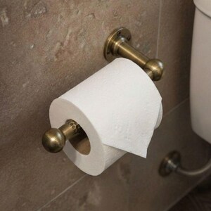 May include: A brass-toned toilet paper holder mounted on a textured, light brown wall. The holder supports a roll of white toilet paper. The design includes decorative ball finials and a curved arm.