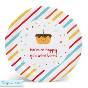 May include: A white round plate with red, yellow, and blue stripes. The center of the plate has a cartoon image of a birthday cake with a single lit candle and the text "We're so happy you were born!"