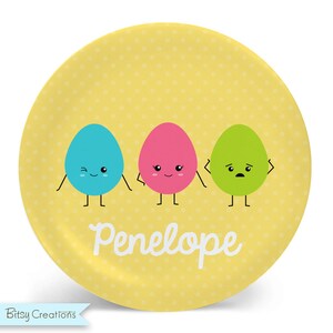 May include: A yellow plate with a polka dot pattern features three cartoon eggs with different facial expressions. The eggs are blue, pink, and green. The plate has the name "Penelope" written in white cursive.