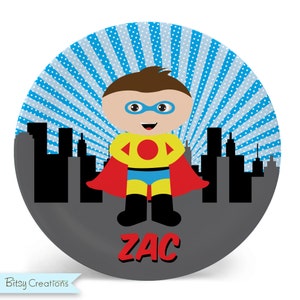 May include: A round, gray and blue plate with a superhero design. A cartoon superhero with a red cape, yellow shirt, and blue pants stands in front of a cityscape. The name "ZAC" is written in red letters at the bottom of the plate.