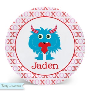 May include: A white round plate with a blue monster holding a red heart. The plate has a pink and red XOXO pattern around the edge. The monster's name is Jaden.