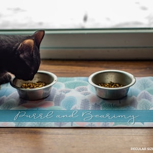 Pode incluir: Um tapete de comida para animais de estima&ccedil;&atilde;o azul e branco com o texto "Purr and Bearimy" nele. Duas tigelas de a&ccedil;o inoxid&aacute;vel est&atilde;o no tapete, uma com comida para gatos. Um gato preto est&aacute; comendo da tigela.